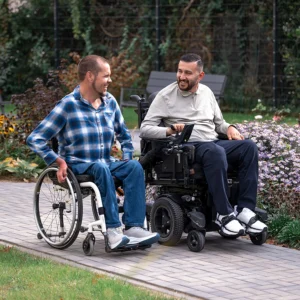 Lars und Mahmoud fahren nebeneinander auf einem Gartenweg im Rollstuhl und lachen sich an.